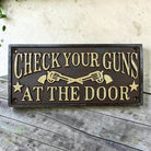 Jones Peak cast iron wall plaque with raised text "Check Your Guns At The Door" and crossed revolvers.